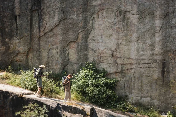 Vue latérale du couple multiethnique senior avec sacs à dos marchant sur le sentier près de la falaise — Photo de stock