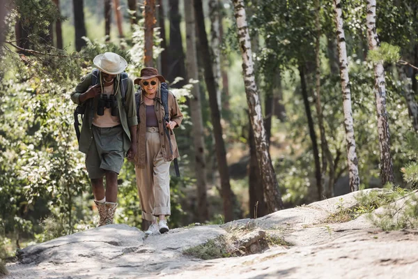 Sorridente donna anziana con zaino che cammina vicino al marito afroamericano nella foresta — Foto stock