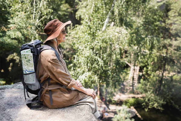 Vista lateral de una anciana con mochila y mapa sentado en el acantilado - foto de stock