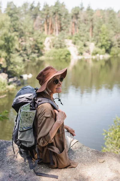 Randonneur sénior joyeux avec sac à dos et carte assis sur une falaise près du lac — Photo de stock