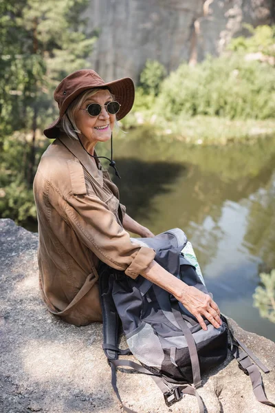 Старший турист в солнечных очках сидит рядом с рюкзаком на скале — стоковое фото