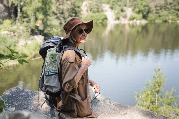 Senderista positivo con mochila y mapa sentado en roca cerca del lago - foto de stock