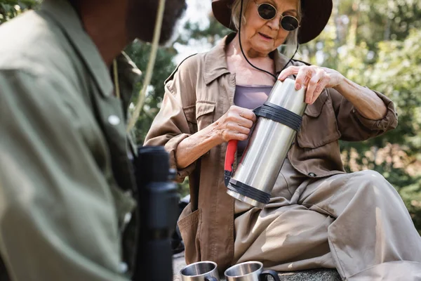 Senior tourist holding thermos near cups and blurred african american husband — Stock Photo