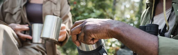 Обрезанный вид на африканских американских туристов, держащих термос возле размытой жены с чашками в лесу, баннер — стоковое фото