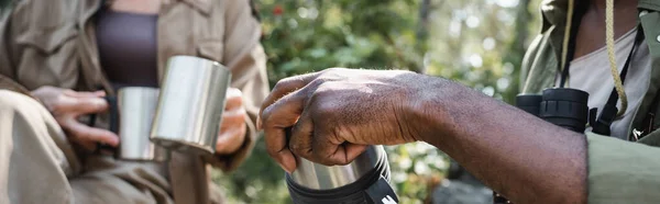 Vista cortada do turista afro-americano segurando garrafa térmica perto da esposa borrada com copos na floresta, bandeira — Fotografia de Stock