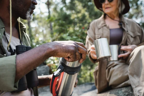 Vista cortada de Africano americano turista segurando thermos perto esposa borrada com copos na floresta — Fotografia de Stock