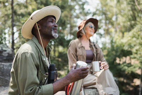 Smiling african american tourist with thermos and binoculars near blurred wife in forest — Stock Photo