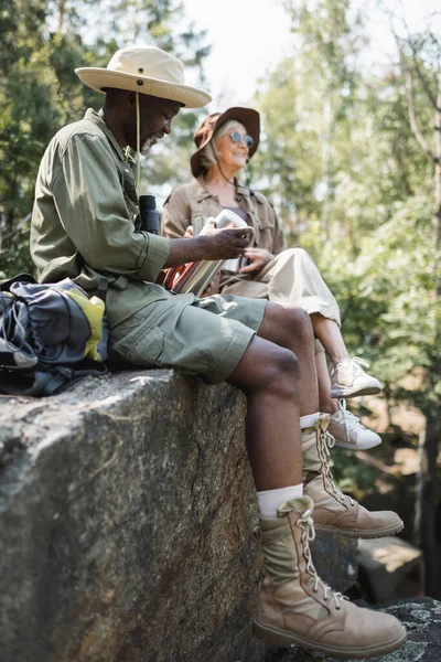 Lächelnder afrikanisch-amerikanischer Wanderer mit Thermoskanne neben Rucksack und verschwommener Frau auf Felsen — Stockfoto
