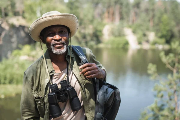African american hiker with backpack and binoculars looking at camera outdoors — Stock Photo