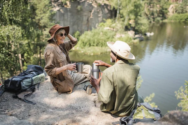 Cheerful senior traveler holding cup near african american husband with thermos on cliff near lake — Stock Photo