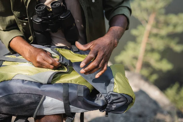 Cropped view of african american tourist with binoculars holding backpack outdoors — Stock Photo