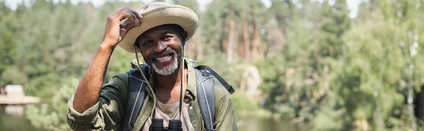 Lächelnder afrikanisch-amerikanischer Tourist mit Hut und Fernglas, der im Freien in die Kamera schaut, Banner — Stockfoto