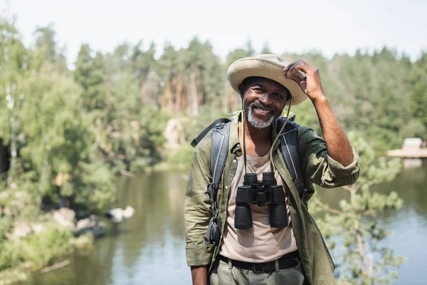 Positive african american hiker with hat and binoculars looking at camera outdoors — Stock Photo