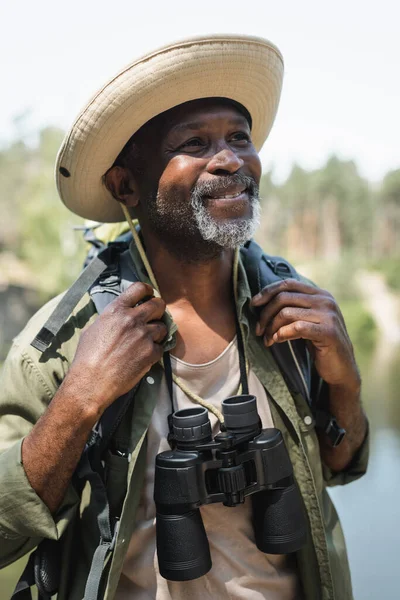 Turista afroamericano feliz con prismáticos y mochila al aire libre - foto de stock