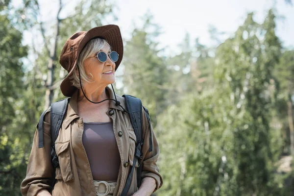 Positiver Wanderer mit Sonnenbrille und Hut steht im Wald — Stockfoto