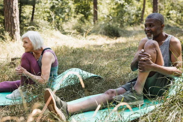 Sorridente uomo africano americano che si estende sul tappeto fitness vicino moglie nella foresta — Foto stock