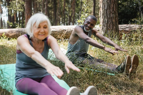 Sonriente afroamericano hombre entrenando en la colchoneta de fitness cerca de la esposa mayor en el bosque - foto de stock