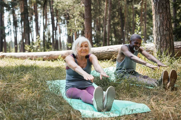 Positiva deportista de edad avanzada haciendo ejercicio en la alfombra de fitness cerca de marido afroamericano en el bosque - foto de stock