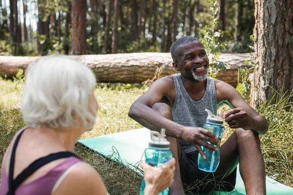 African american sportsman holding sports bottle near blurred wife in forest — Stock Photo