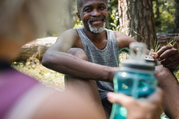 Esportista afro-americano sorridente segurando garrafa de esportes perto da esposa borrada na floresta — Fotografia de Stock