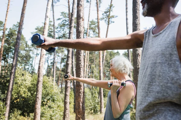 Vista laterale della donna anziana che si allena con i manubri vicino al marito afroamericano offuscato nella foresta — Foto stock