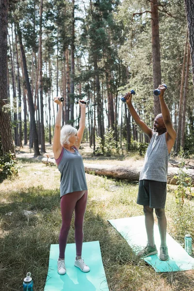 Cheerful interracial couple training with dumbbells on fitness mats in forest — Stock Photo