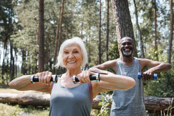 Sonriente anciana haciendo ejercicio con mancuernas cerca borrosa afroamericano marido en el bosque - foto de stock