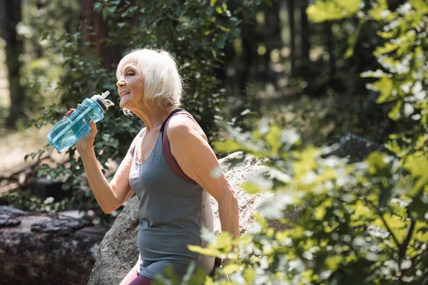 Sorridente sportiva anziana che tiene una bottiglia di sport con acqua vicino alla pietra nella foresta — Foto stock
