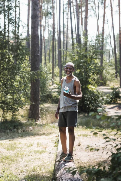 Sorridente sportivo africano americano che tiene una bottiglia sportiva sul tronco nella foresta — Foto stock