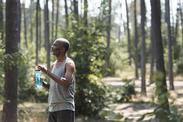 Side view of elderly african american sportsman holding sports bottle in forest — Stock Photo