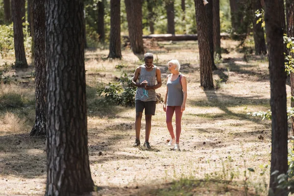 Senior pareja interracial con botella deportiva caminando en el bosque de verano - foto de stock