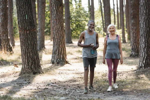 Elderly african american sportsman with sports bottle walking near smiling wife in forest — Stock Photo