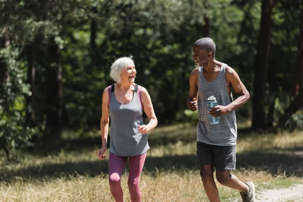 Cheerful multiethnic couple with bottle of water running in forest — Stock Photo