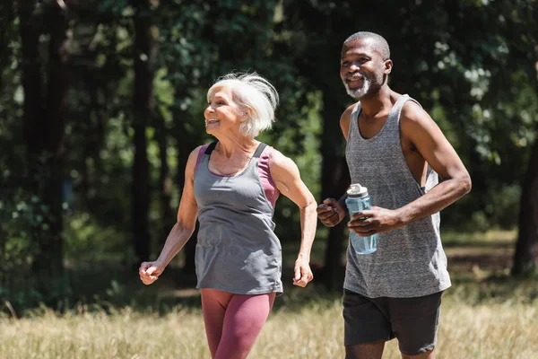 Hombre afroamericano mayor con botella deportiva corriendo cerca de esposa sonriente en el bosque - foto de stock