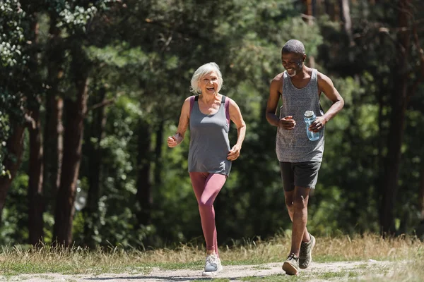 Cheerful multicultural couple with sports bottle running in forest — Stock Photo