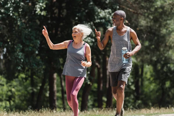 Happy multiethnic couple with sports bottle pointing with fingers while running in park — Stock Photo