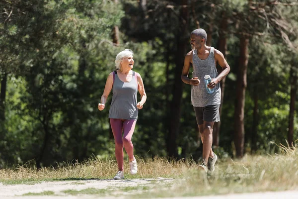 Afro-americano deportista corriendo cerca alegre senior esposa en bosque - foto de stock
