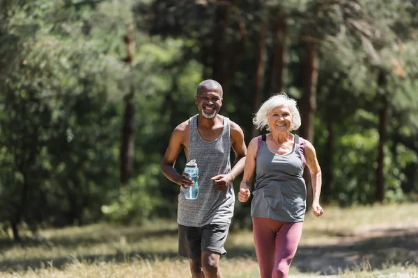 Smiling elderly sportswoman running near african american husband with sports bottle in forest — Stock Photo