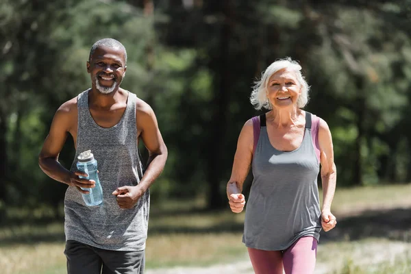 Cheerful interracial couple with sports bottle jogging and looking at camera outdoors — Stock Photo