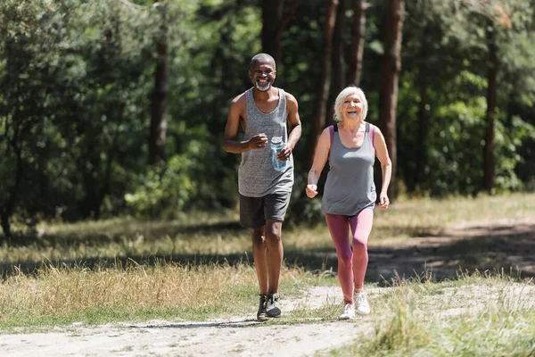 African american sportsman with sports bottle running near happy wife in forest — Stock Photo