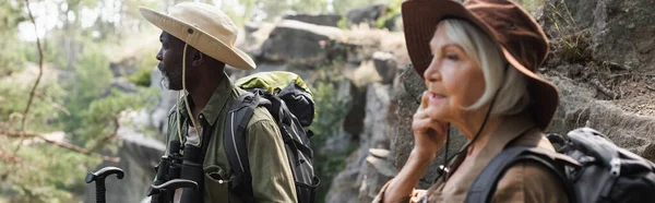 Senior african american tourist with trekking poles standing near blurred wife in forest, banner — Stock Photo