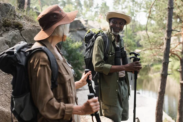 Lächelnder afrikanisch-amerikanischer Tourist mit Trekkingstöcken blickt verschwommene Frau im Wald an — Stockfoto