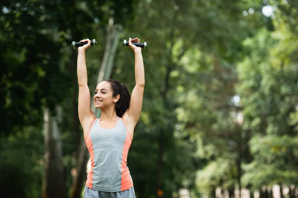 Cheerful sportswoman holding dumbbells above head in park — Stock Photo