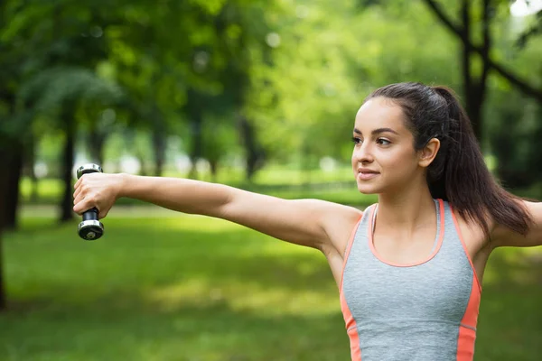 Young sportswoman smiling while exercising with dumbbell in park — Stock Photo