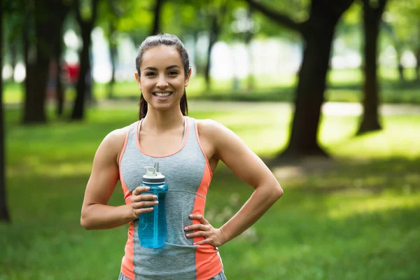 Donna in forma allegra che tiene bottiglia sportiva mentre in piedi con mano sul fianco nel parco — Foto stock