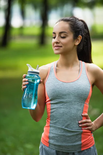 Happy and fit woman holding sports bottle while standing with hand on hip — Stock Photo