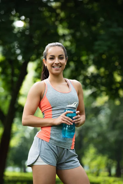 Счастливая и здоровая женщина, держа в руках спортивную бутылку, стоя в парке — стоковое фото
