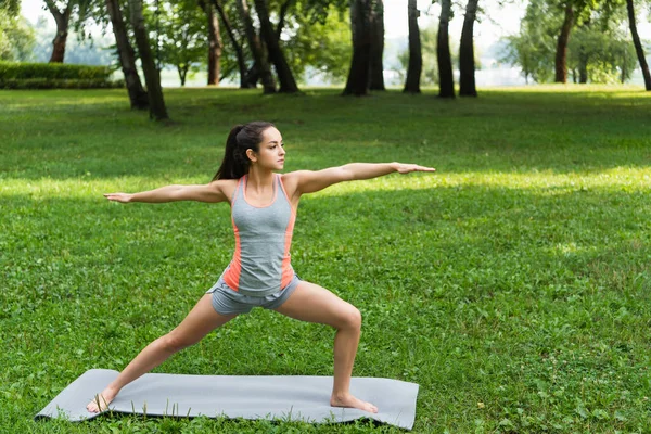 Giovane donna in piedi in posa guerriero sul tappeto yoga nel parco — Foto stock