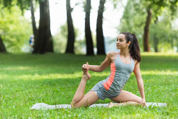 Flexible woman in sportswear stretching on yoga mat in park — Stock Photo