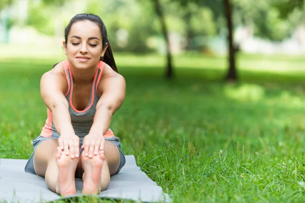 Гибкая и улыбающаяся женщина в спортивной одежде, растянутая на коврике для йоги в парке — стоковое фото